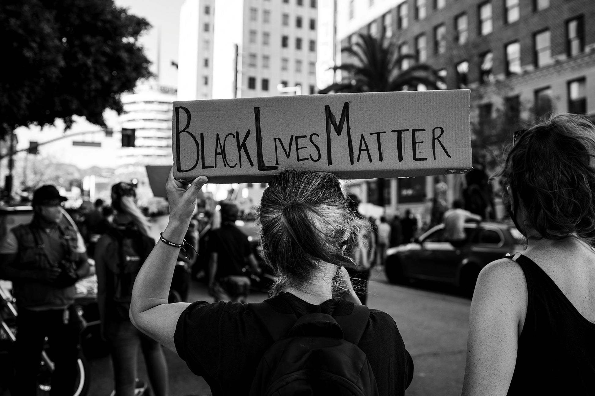 "Advocate" is a black and white fine art photograph captured during the Black Lives Matter March in Hollywood, California, on June 7, 2020. Shot from behind, the image portrays a woman with her head held high, adorned with a Black Lives Matter cardboard sign.  By Shannon Black