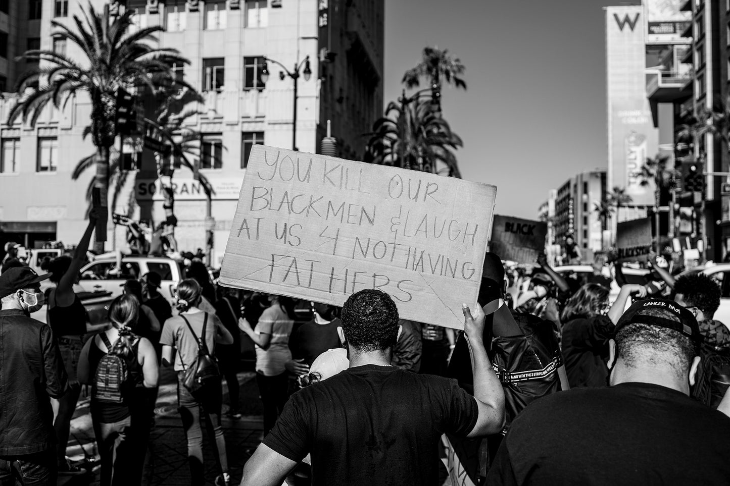 Shot from behind, the image portrays a man standing amidst a sea of people, his raised Black Lives Matter cardboard sign bearing a powerful message: "You kill our Black Men & laugh at us for not having fathers." 